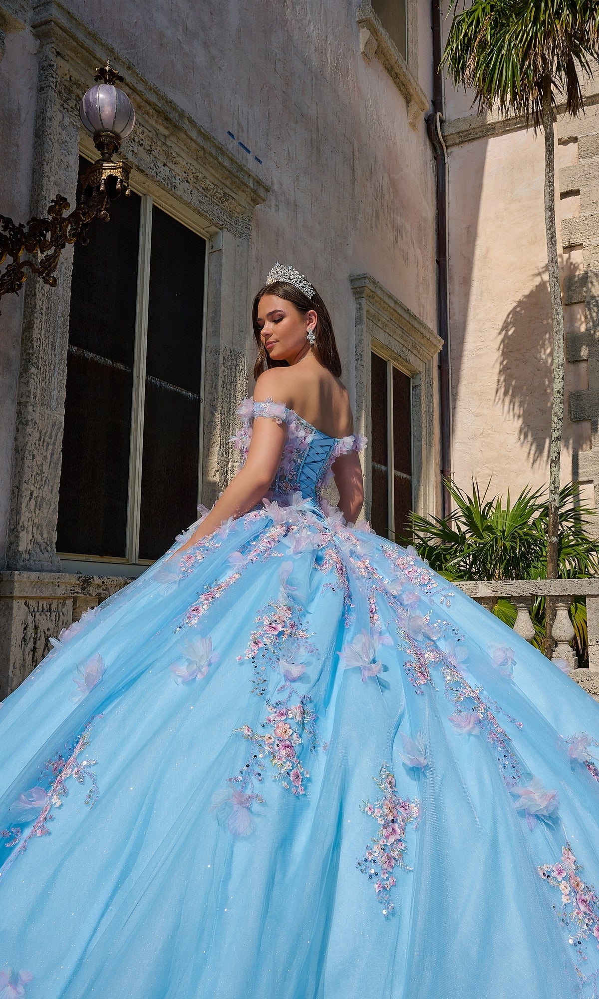 A young woman wearing the Quinceanera Dress 54228 By Amarra, an off-the-shoulder blue ball gown with 3D floral appliqué and a tiara, stands outside a stone building, looking over her shoulder.